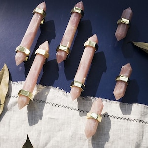 Può includere: Bacchette di cristallo di quarzo rosa con fasce metalliche color oro. I cristalli rosa sono esposti su una superficie blu scuro con un tessuto bianco e foglie color oro. Le bacchette sono appuntite ad entrambe le estremità.