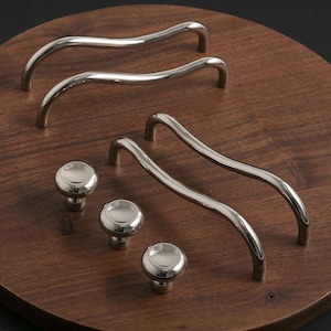 May include: A selection of polished silver cabinet handles and knobs displayed on a dark wooden surface. The curved handles and round knobs are designed for furniture. The hardware reflects light, and is made of a shiny metal.