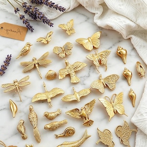 Puede incluir: Una colección de herrajes decorativos dorados con varios diseños inspirados en la naturaleza. El surtido incluye mariposas, pájaros, libélulas, conchas marinas y peces. El hardware está dispuesto sobre una superficie de mármol con ramitas de lavanda y un paño blanco.