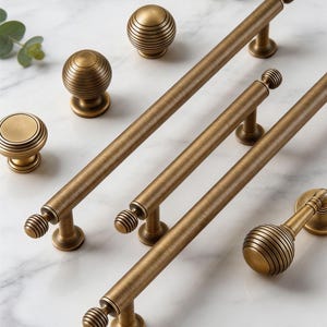 May include: A collection of antique brass cabinet hardware, including knobs and pulls. The knobs have a round, ribbed design, while the pulls are long and cylindrical with similar ribbed detailing on the ends. The hardware is arranged on a white marble surface.