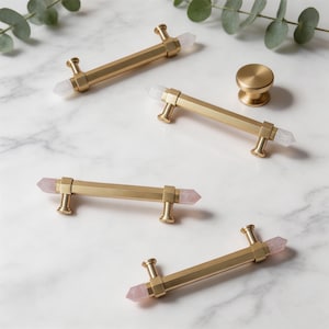 May include: A collection of gold-toned cabinet hardware, including several bar pulls and a single knob. The pulls feature faceted designs and are accented with clear and pink crystal points. The hardware is arranged on a white marble surface.