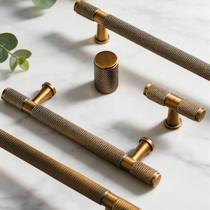 Op de afbeelding: Bronskleurige meubelbeslagset. De set bevat verschillende T-bar handgrepen en een cilindrische knop, allemaal met een textuur, diamantpatroon. De hardware wordt weergegeven op een wit marmeren oppervlak, met groene bladeren op de achtergrond.