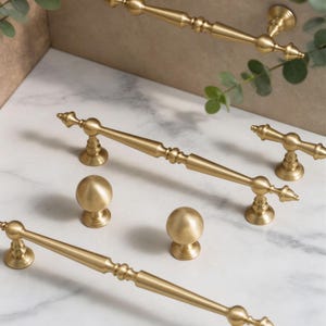 May include: A collection of gold-toned cabinet hardware, including handles and knobs. The handles have a detailed, ornate design with decorative ends. The knobs are spherical. The hardware is arranged on a marble surface with greenery.