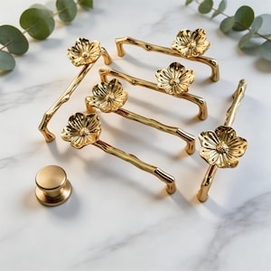 May include: Gold-colored cabinet hardware set featuring floral and branch designs. The set includes several pull handles and a round knob. The handles have a branch-like shape with a flower at the center. The hardware is displayed on a white marble surface.