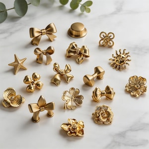 May include: A collection of gold-colored cabinet knobs and pulls in various designs, including bows, flowers, a starfish, and a sun. The hardware is arranged on a white marble surface, showcasing a range of decorative options for furniture and home decor.