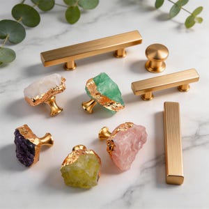 May include: A collection of cabinet hardware, including gold-toned bar pulls, a round knob, and gemstone knobs. The gemstone knobs feature raw, colorful stones like amethyst, rose quartz, and green fluorite, with gold-colored edges. The hardware is arranged on a white marble surface.