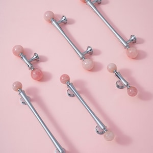 May include: Six cabinet pulls with a polished silver finish and pink, spherical accents. The pulls are arranged on a light pink background. Each pull has a central bar with a sphere at each end, and decorative mounting hardware.