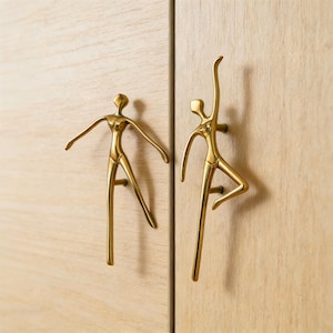 May include: Two brass cabinet handles shaped like stylized human figures. One has outstretched arms, the other is in a ballet pose. They are mounted on light-coloured wooden cabinet doors.