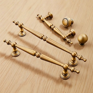 May include: A selection of antique brass cabinet hardware, comprising two long handles, two smaller handles, and two knobs. The hardware features an ornate design and a warm, golden finish. The pieces are displayed on a light wood surface.