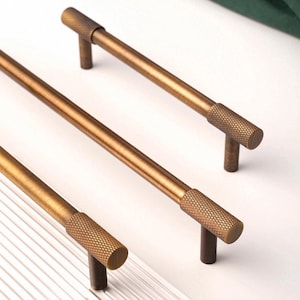 May include: Three bronze-coloured cabinet pulls are displayed. Each pull features a textured cylindrical end and a straight bar. The pulls are arranged diagonally, highlighting their design and finish. Suitable for kitchen or bathroom cabinets.