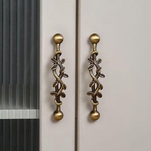 以下が含まれることがあります： 植物のつるのデザインが施された、2つのアンティーク真鍮製キャビネットプル。各プルは、葉と丸みを帯びた端部を備えた中央のつるモチーフが特徴です。プルは、明るい灰色のキャビネットドアに取り付けられています。