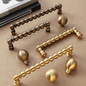 May include: A selection of antique brass cabinet handles and knobs. The handles have a beaded design, and the knobs are spherical. The hardware is presented on a neutral background, highlighting the warm tones of the metal. The handles are approximately 15 cm long.