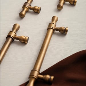 May include: Four antique brass cabinet pulls are displayed. Each pull has a T-shaped design with a cylindrical handle and a decorative base. The brass finish has a slightly aged appearance. The pulls are arranged on a light-coloured surface.