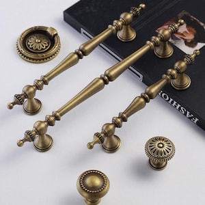 May include: A selection of antique brass cabinet hardware, including handles, knobs, and a pull ring. The hardware features ornate floral and geometric designs, with a warm, aged finish. The handles are approximately 15 cm long.