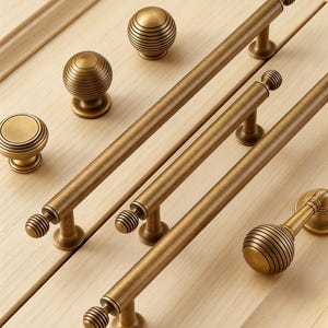 May include: Close-up of various antique brass cabinet hardware, including knobs and pulls. The hardware features a warm, aged finish and a ribbed design. The pulls are long and straight, while the knobs are round.