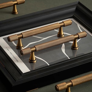 May include: Close-up of several antique brass cabinet pulls with a textured, diamond-patterned design. The T-bar handles are mounted on a black framed artwork with a grey and white abstract design. The hardware is displayed on a dark green surface.