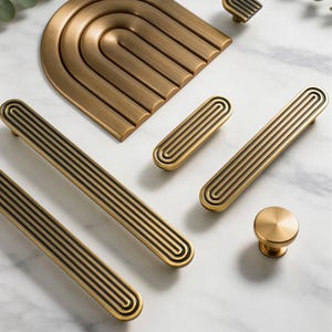 May include: A collection of brass-toned cabinet hardware, including handles, knobs, and a decorative arch. The hardware features a grooved design with black accents. The pieces are arranged on a white marble surface.