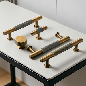 May include: A collection of brass cabinet hardware, including handles and a knob, arranged on a white surface. The handles have a ribbed, dark gray center section. The hardware is designed with a vintage aesthetic.