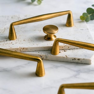 May include: A collection of gold-toned cabinet hardware, including several bar pulls and a round knob. The hardware is arranged on a light-colored, textured surface, with green foliage in the background. The hardware has a brushed finish.