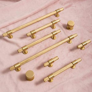 May include: A selection of gold-coloured cabinet hardware, including bar pulls and round knobs. The hardware has a textured, diamond-patterned design. The items are arranged on a pink fabric surface, showcasing their metallic finish and design.
