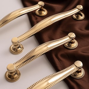 May include: Four gold cabinet handles with a classic design. Each handle has a curved bar with a beaded edge and a circular base. The handles are displayed on a white and brown background.