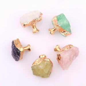 May include: Five cabinet knobs with natural stone faces in white, green, purple, yellow-green, and pink. Each stone is edged with gold and mounted on a gold-coloured base. These knobs add a unique, natural touch to furniture.