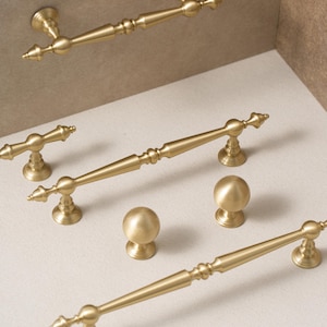 May include: A selection of gold-coloured cabinet hardware, including bar pulls and round knobs. The pulls have a decorative, detailed design with pointed ends. The knobs are spherical. The hardware is displayed against a neutral background.