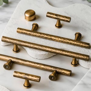 May include: A collection of antique brass cabinet hardware, including bar pulls, T-shaped pulls, and a round knob. The hardware has a hammered texture and a warm, golden hue. The items are arranged on a white surface with a marble-like appearance.