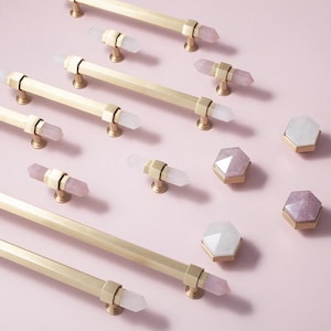 May include: A collection of gold-coloured cabinet pulls and knobs, each featuring crystal embellishments. The pulls are long and rectangular, while the knobs are hexagonal. Some crystals are clear, others are pink, set on a pale pink surface.