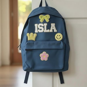 Peut inclure: Un sac à dos bleu marine avec le nom "ISLA" en lettres blanches. Le sac à dos est décoré d'un nœud jaune, d'une couronne, d'un smiley et d'une fleur rose. Le sac à dos est accroché à une porte blanche.