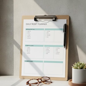 May include: A clipboard with a "DAILY RESET PLANNER" sheet, a black pen, and a small potted succulent. The planner has sections for morning routine, afternoon tasks, and evening rituals. Brown-framed glasses rest below the clipboard.