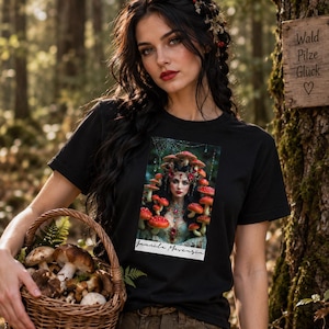 Könnte beinhalten: Ein schwarzes Kurzarm-T-Shirt mit einem rechteckigen Grafikdruck einer Person, die mit roten Pilzen und einer dekorativen Halskette geschmückt ist. Der Druck enthält den kursiven Text "Jannila Merenzia" vor einem dunklen Waldhintergrund.