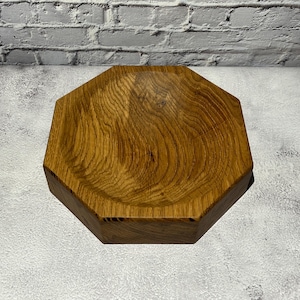 May include: An octagonal wooden bowl with a natural wood grain finish. The bowl is made of light brown wood and has a shallow, concave interior. The bowl is displayed on a light grey surface.