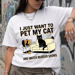 Könnte beinhalten: Weißes T-Shirt mit dem Text "I JUST WANT TO PET MY CAT AND WATCH MURDER SHOWS". Die Grafik zeigt eine schwarze Katze auf einem gelben Sofa, einen Fernseher mit einem Detektiv und einen kleinen Tisch. Das Shirt ist ein Freizeitartikel.