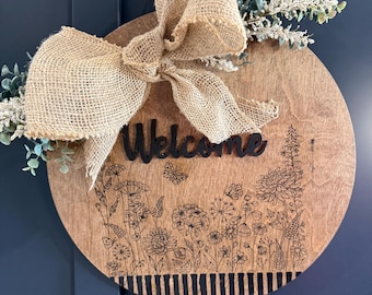 May include: A round wooden welcome sign with a burlap bow and greenery. The sign features a floral design and the word "Welcome" in black. The sign is approximately 12 inches in diameter.