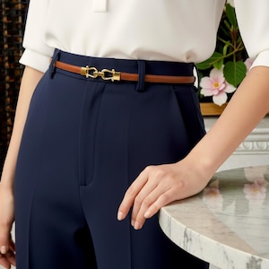 May include: A brown leather belt with a gold-colored buckle is worn with navy blue pants. The belt has a thin profile and a classic design. The buckle is a decorative element, adding a touch of elegance to the outfit. The pants are high-waisted.