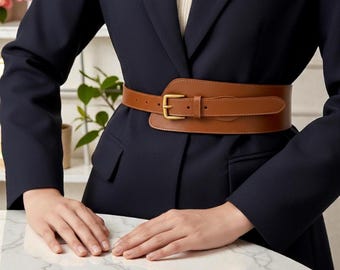May include: A wide, brown leather belt with a gold-colored buckle is worn over a navy blue blazer. The belt has a rectangular design with a rounded edge and a classic buckle closure. The blazer has a tailored fit.