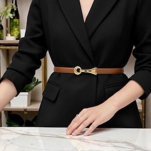 Puede incluir: Un cinturón de cuero marrón con una hebilla dorada se lleva sobre una blazer negra. El cinturón tiene un diseño sencillo y elegante, con una correa fina y una hebilla decorativa. La blazer tiene un corte clásico.