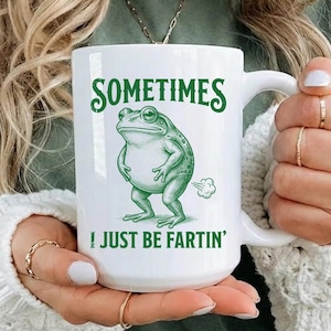 May include: White ceramic mug with a green frog illustration and the text "SOMETIMES I JUST BE FARTIN'". The frog is depicted with a humorous expression. The mug has a large handle.