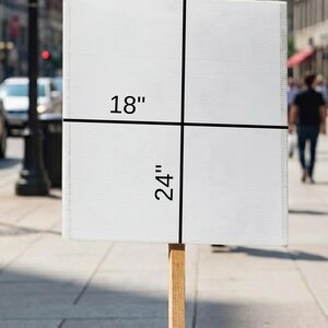 May include: A white rectangular sign, 18 inches by 24 inches, mounted on a wooden pole. The sign is blank, with black lines indicating the dimensions. The background shows a city street with pedestrians and buildings.