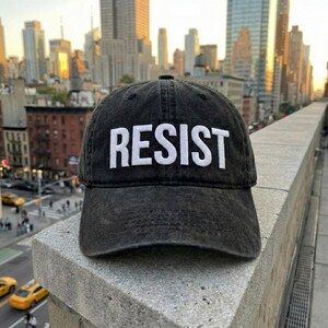 May include: Black, distressed baseball cap with the word "RESIST" embroidered in white block letters. The cap is set against a cityscape background with tall buildings and a bright sky.