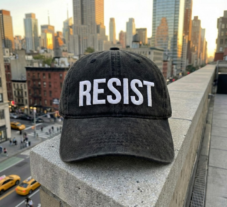 May include: Black, distressed baseball cap with the word "RESIST" embroidered in white block letters. The cap is set against a cityscape background with tall buildings and a bright sky.