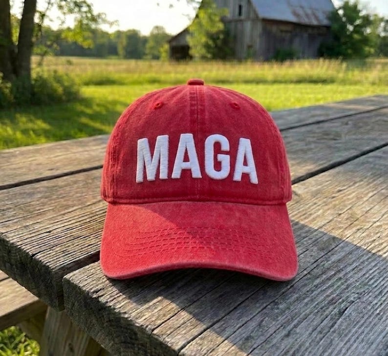 May include: A red baseball cap with a distressed, washed-out texture featuring the word MAGA embroidered in bold, white, capital letters across the front panel. The cap has a curved brim and a structured crown with visible stitching details.
