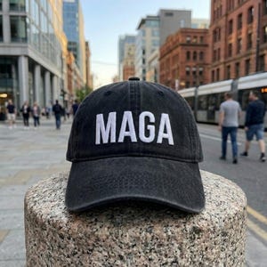 May include: A black baseball cap with a washed, distressed texture featuring the text MAGA embroidered in white capital letters across the front panel. The cap has a curved brim and a structured crown, showcasing a classic six-panel design.