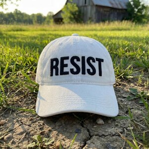 May include: White baseball cap with the word "RESIST" embroidered in bold black letters. The cap is sitting on the ground in a grassy field. The hat has a curved brim and a classic six-panel design.
