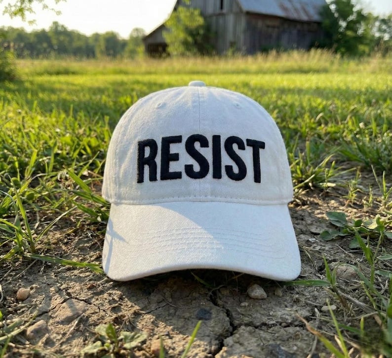 May include: White baseball cap with the word "RESIST" embroidered in bold black letters. The cap is sitting on the ground in a grassy field. The hat has a curved brim and a classic six-panel design.