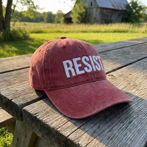 May include: A red baseball cap with the word "RESIST" embroidered in white block letters. The cap is made of a soft, worn material and sits on a weathered wooden picnic table. The background features a rural landscape.