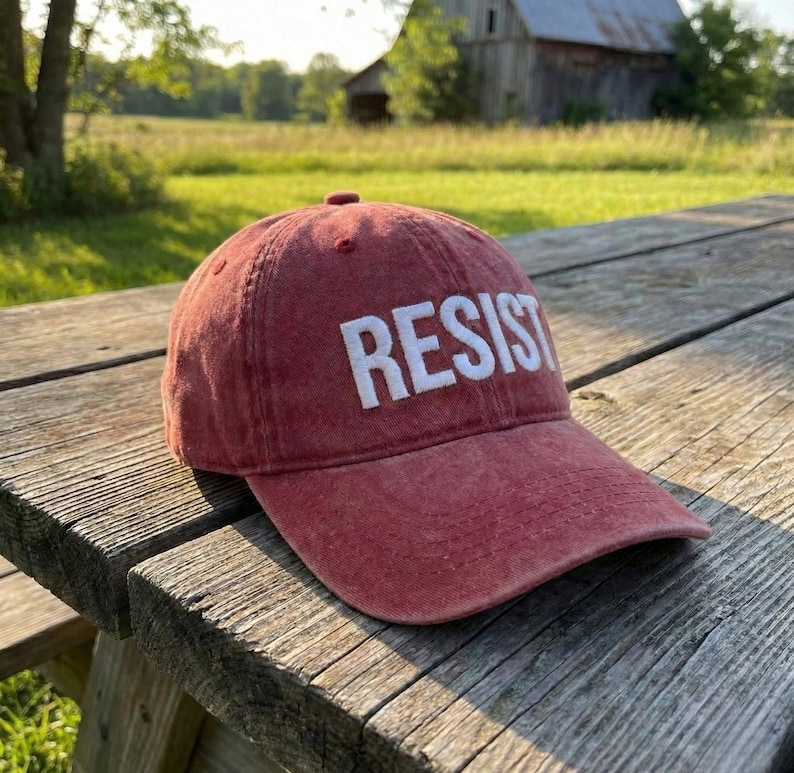 May include: A red baseball cap with the word "RESIST" embroidered in white block letters. The cap is made of a soft, worn material and sits on a weathered wooden picnic table. The background features a rural landscape.