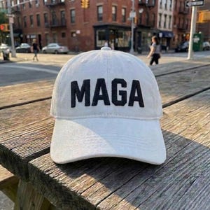 May include: A white baseball cap featuring the word MAGA embroidered in bold black capital letters across the front panel. The cap has a structured crown, a curved brim with visible stitching, and a classic six-panel design with a button on top.