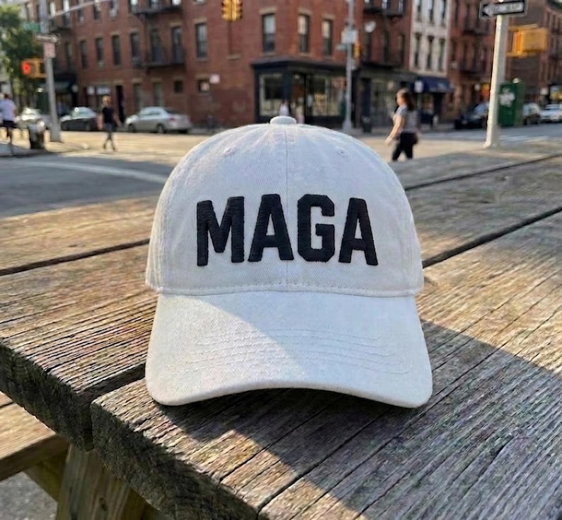 May include: A white baseball cap featuring the word MAGA embroidered in bold black capital letters across the front panel. The cap has a structured crown, a curved brim with visible stitching, and a classic six-panel design with a button on top.